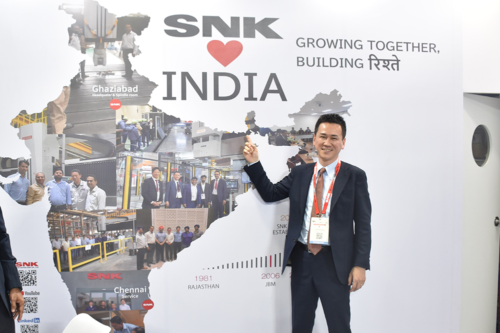 SNK President Akira Nakanishi standing before a booth decorated to highlight the company’s strong ties with India. 